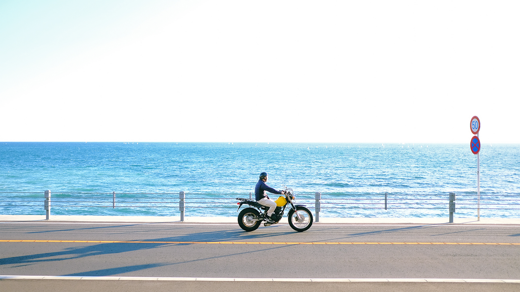 Man riding yellow motorcycle on a road along the beach with blue ocean water and horizon in the background.