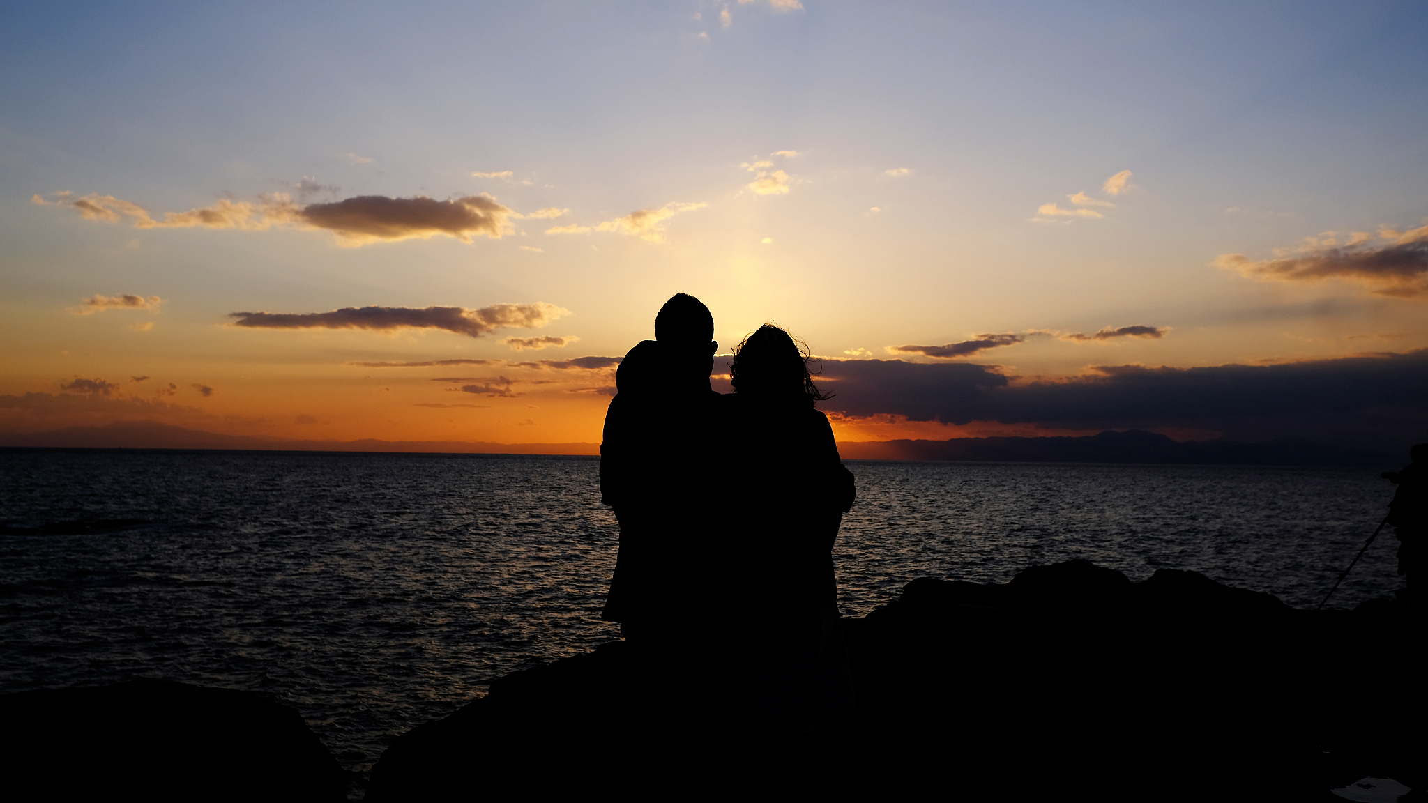 Silhouette of a couple standing on ocean rocks in the sunset.